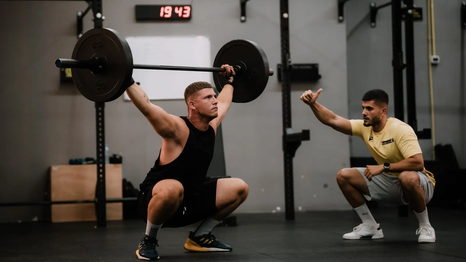 Lifter holding a barbell overhead at the bottom of a squat while his coach squats beside him and gives a thumbs-up